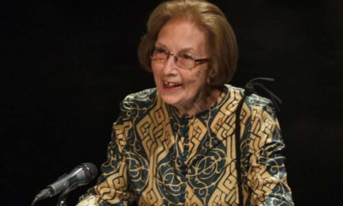 Elderly person stood at a podium giving a speech. They are wearing a patterned blouse and glasses.