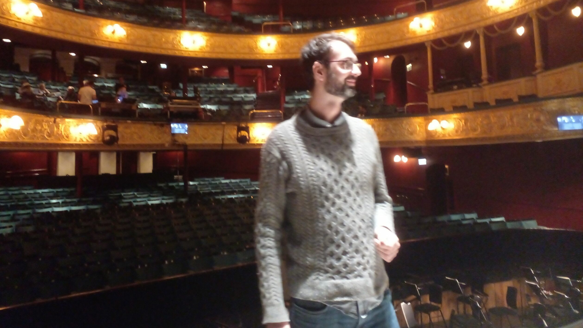 Person stood on stage looking toward the back of the stage, the lights are illuminating the back of the theatre