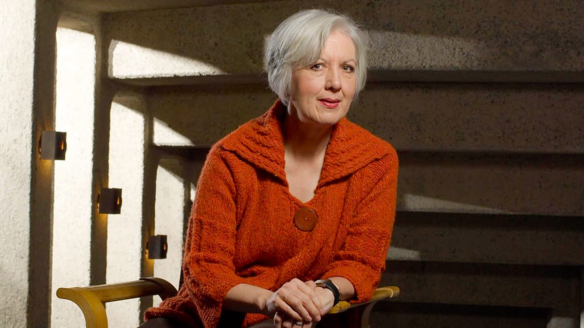 Judith Weir sat on a wooden chair, they have their hands in front of them.