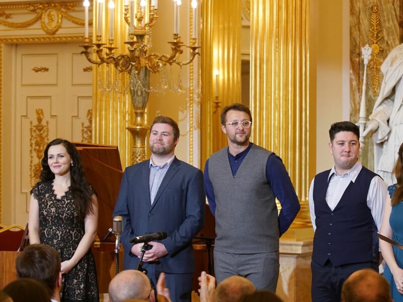 Group of people stood in a grand room being interviewed by someone with a microphone on the right hand side