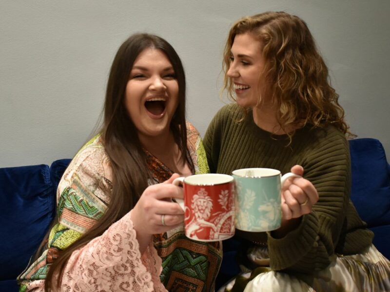 Two people sat on a sofa, they both are cheers-ing their mugs and have their mouths open wide smiling