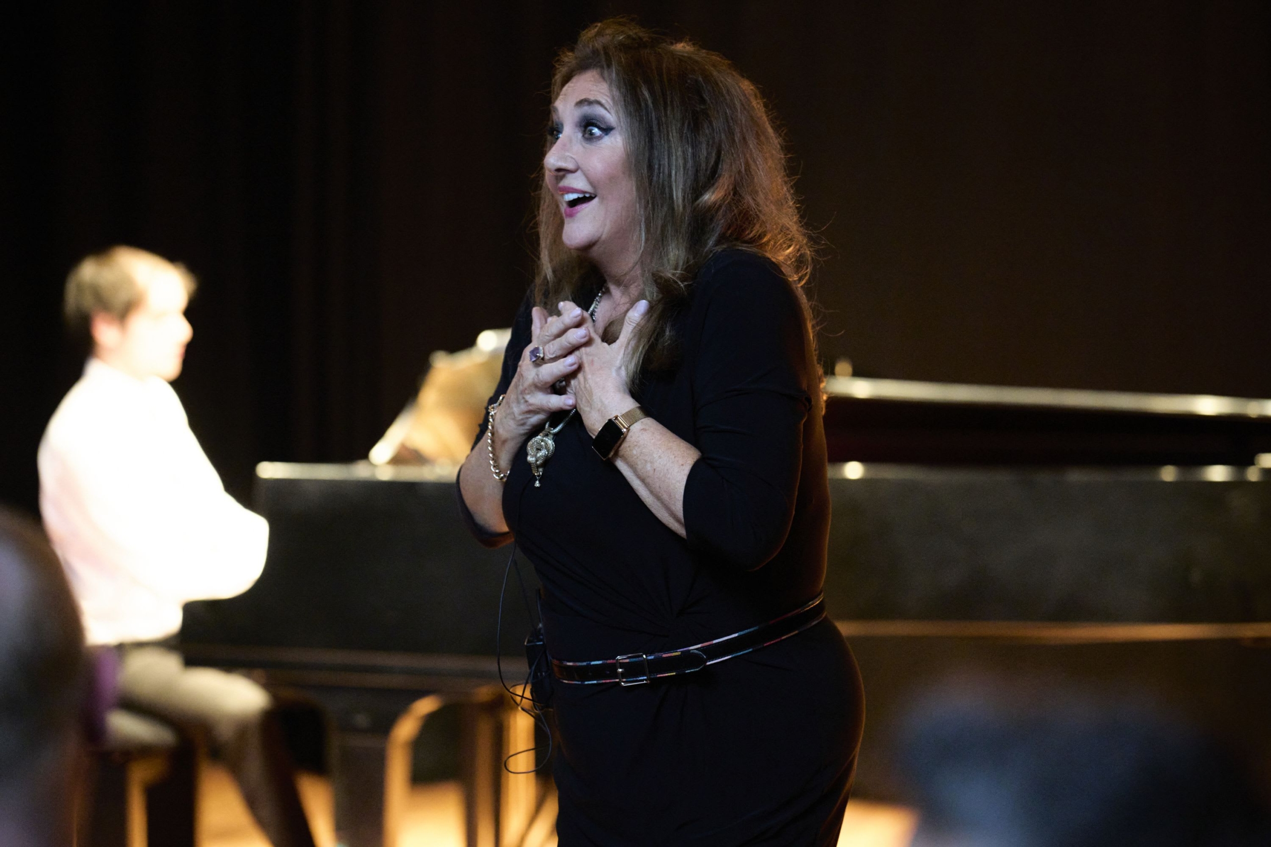 Person on stage holding their hands up to their chest, they look elated. There is someone in the background playing piano.