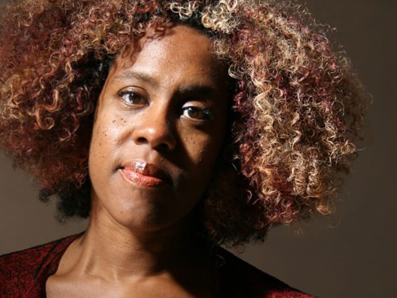 Portrait of Errollyn Wallen. They have curly short hair and are smiling at the camera,