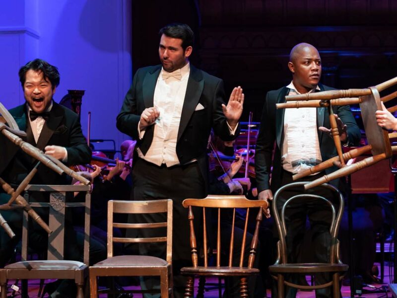 Group of people on stage, they are all stood in front of a chair. People on each side are holding up the chairs to each other.