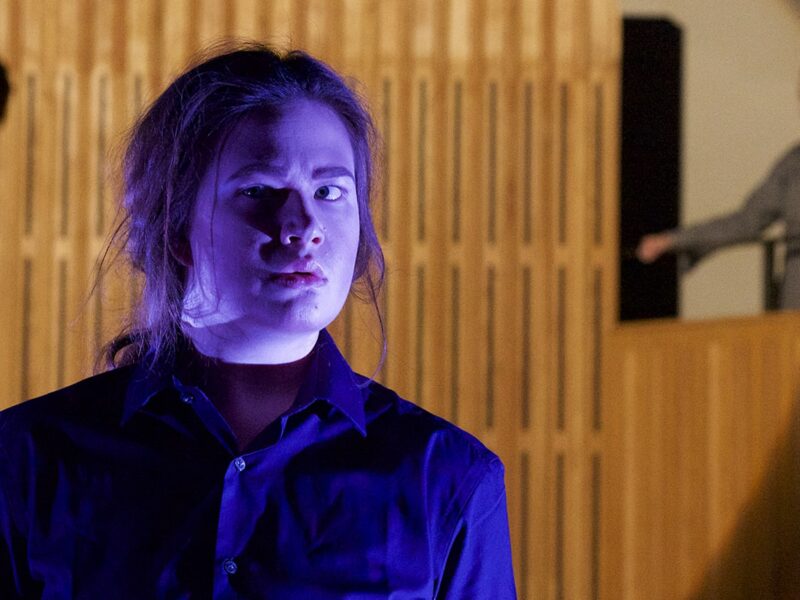 Person with long hair tied back performing on stage, they are wearing a blue shirt and are illuminated by purple lighting. There are two people stood on a podium behind.