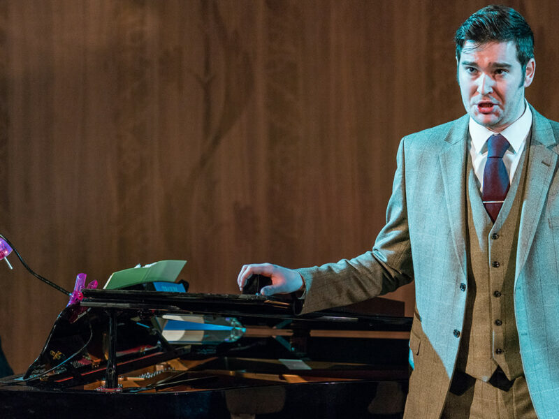 Person performing on stage wearing a suit, they have their arm resting on a piano where two people are sat playing