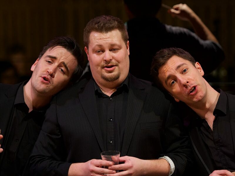 Three people on stage wearing black suits. There is someone in the middle and two either side resting their head on the middle persons shoulder