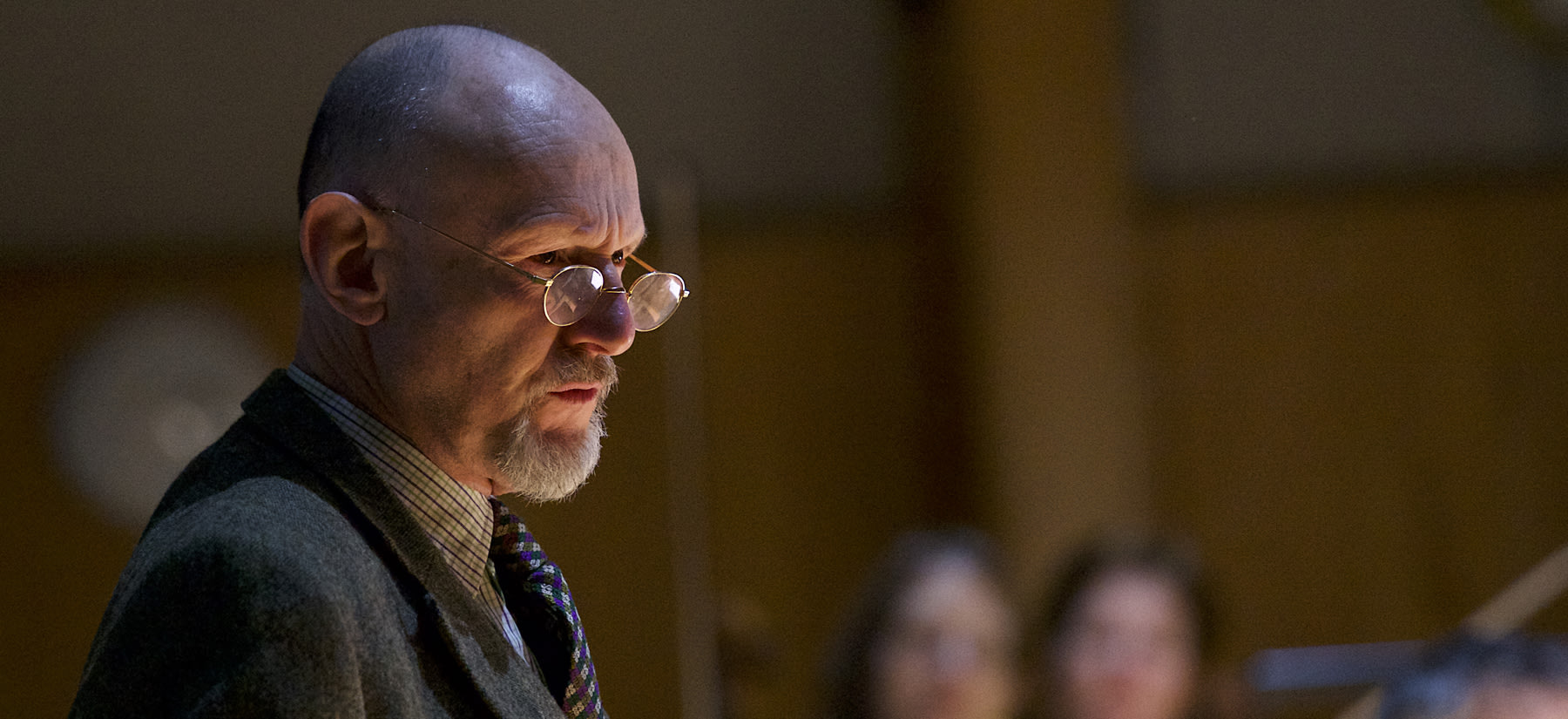 Person wearing glasses and a suit with a very stoic expression, they are looking to the side.