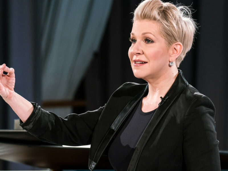 Person with short blonde hair stood next to a piano, they are holding their arm up in the air looking to the side