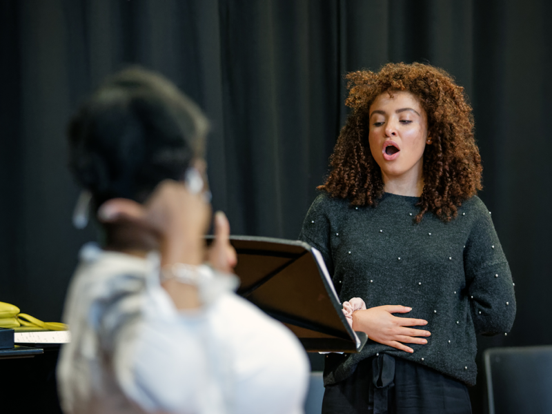 Person holding their hand to their stomach singing, stood in front of a music stand