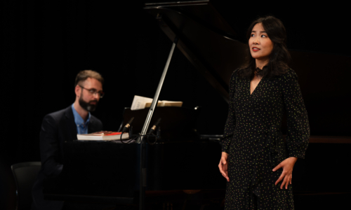 Person performing on stage with another person behind them playing piano