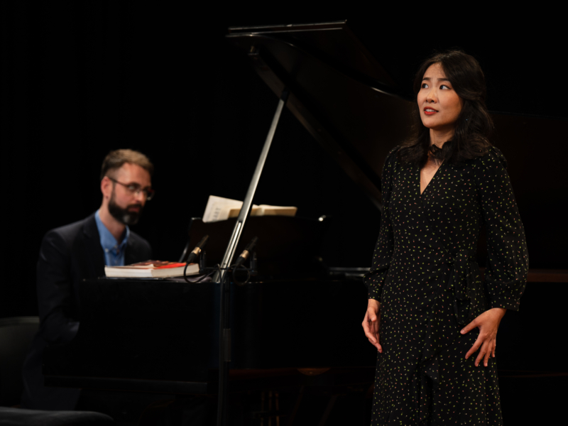 Person performing on stage with another person behind them playing piano