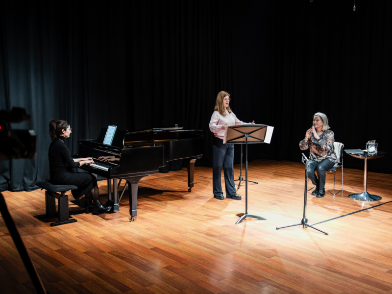 Two people in a room performing, one person is sat to the right watching