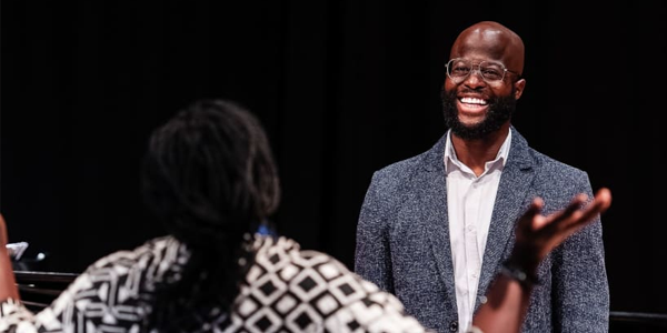 Person smiling widely, wearing smart clothing and glasses. Someone is wearing a patterned top with their back to the camera with their arms up in the air.