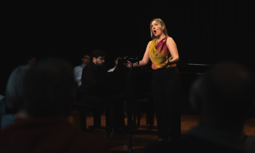 Carmen Lazaro performs in a pink-yellow top in front of a black piano and backdrop, singing in a Wandsworth Wednesday free lunchtime recital