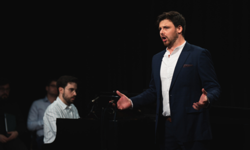 Ambrose Connolly in a white shirt and blue suit performs in front of a black piano and background in a Wandsworth Wednesday free lunchtime recital