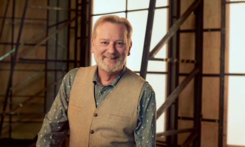 David Gowland poses smiling. He's backstage with the backs of scenery for a backdrop