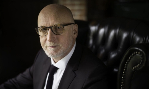 Brindley Sherratt in a black suits sits on a black leather chair